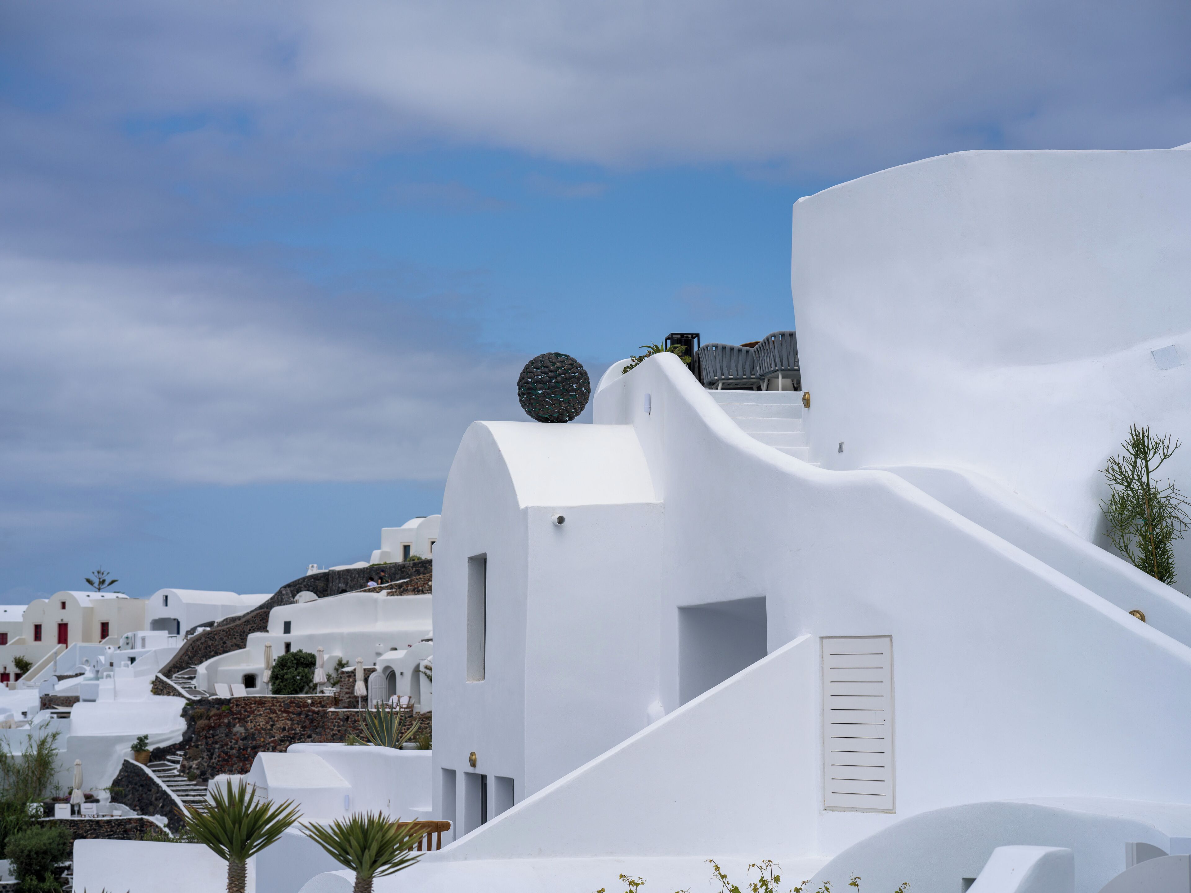 A white-walled building with a staircase leading to a door, surrounded by palm trees and other white buildings.