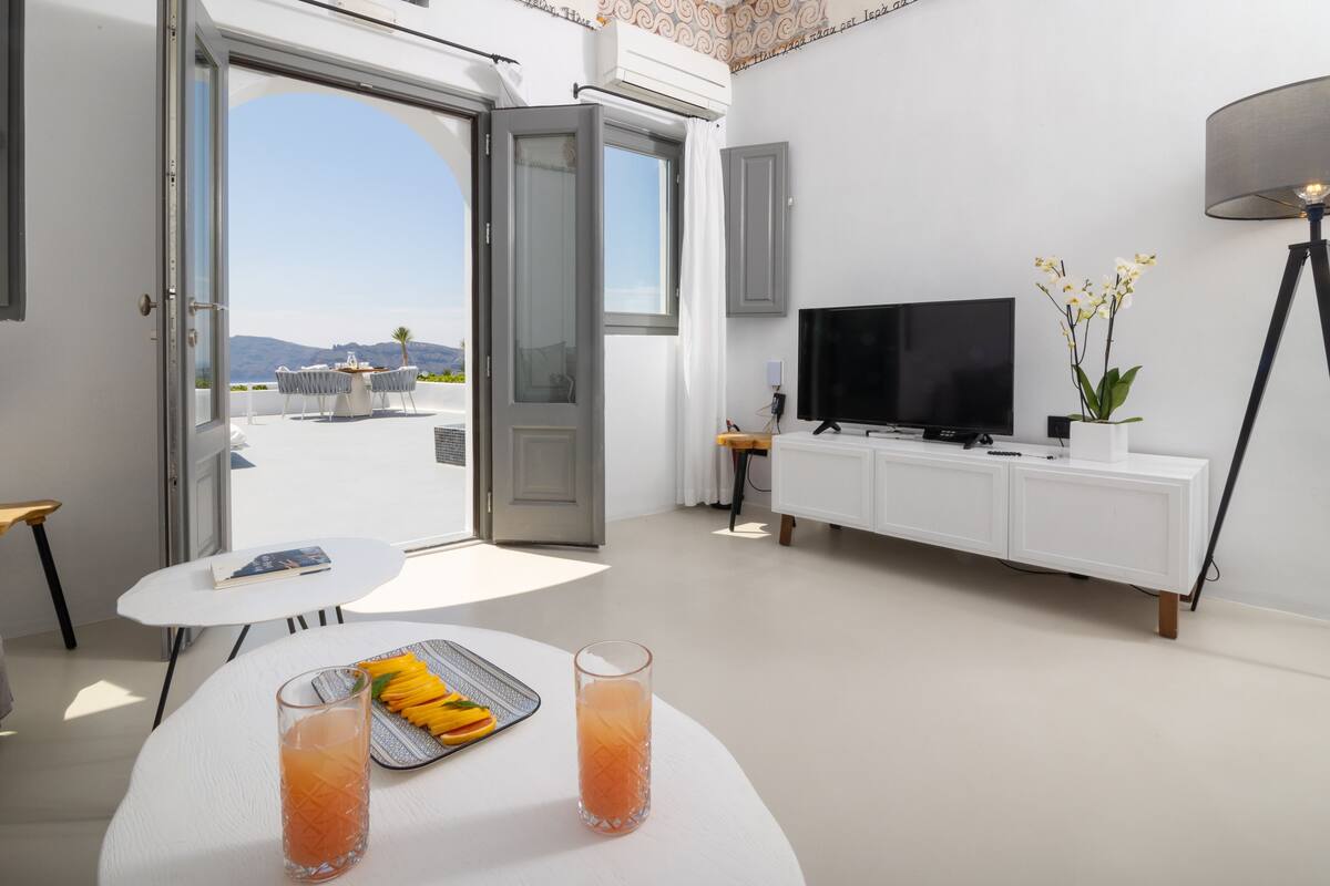 A modern living room with a large window, a flat-screen TV, a dining table with fruit and drinks, and a white cabinet.