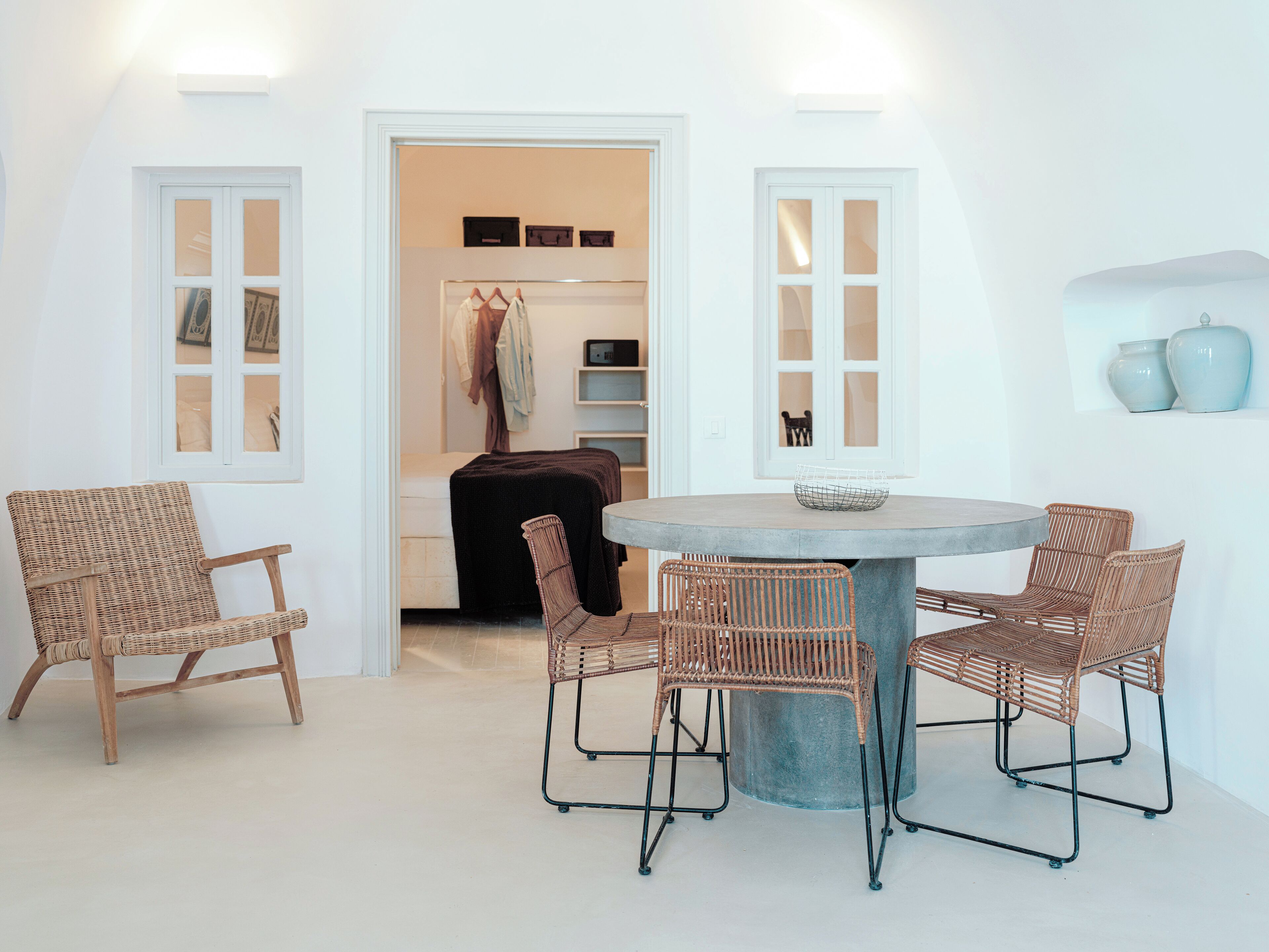A dining area with a round table and four wicker chairs, a bench, and a door leading to a room with a coat rack and shelves.