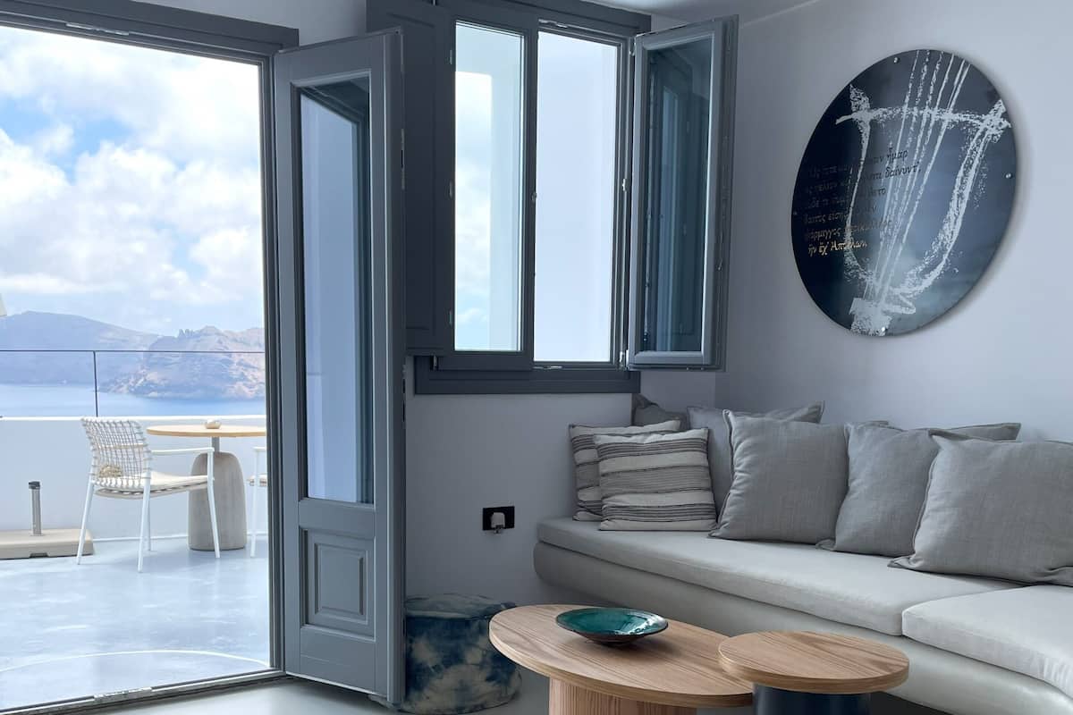 A modern living room with a sofa, a round wooden coffee table with a bowl, a large window with a view of the sea, and a wall-mounted abstract art piece.