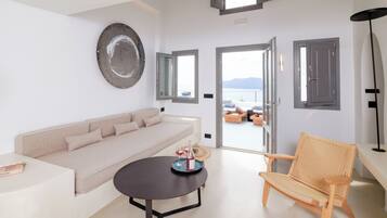A modern living room with a sofa, a round table with a bottle and glasses, a wicker chair, and a view of the sea through open doors.
