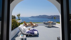 A terrace with a view of the sea, featuring lounge chairs, a table, and a white wall. - IKIES Santorini (Santorini)