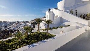 A white-washed building with a curved roof and a small dome, surrounded by greenery and palm trees. - IKIES Santorini (Santorini)
