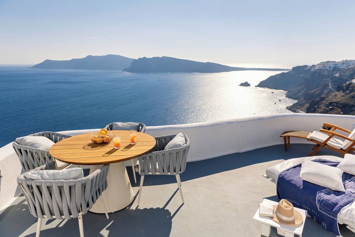 A terrace with a round wooden table, two grey upholstered chairs, a lounge chair, and a bed with a blue cover.