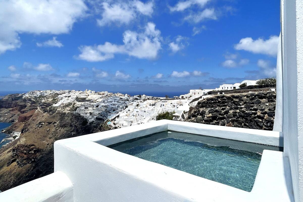A white building with a pool, overlooking a coastal village with white houses and rocky terrain.