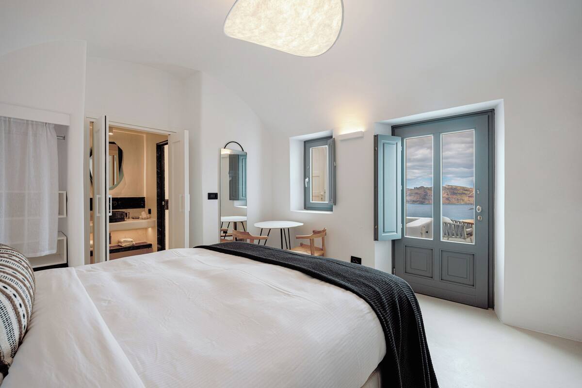 A hotel room with a large bed, a small table, and a view of the sea through a door.