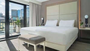 Una habitación de hotel moderna con una cama grande, un pequeño banco, una mesita de noche con lámpara y vistas de la ciudad a través de amplios ventanales.