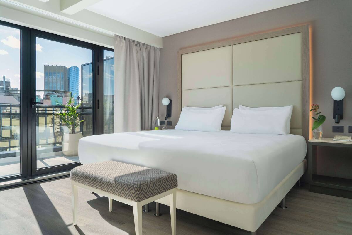 A modern hotel room with a large bed, a small bench, a bedside table with a lamp, and a view of the city through large windows.