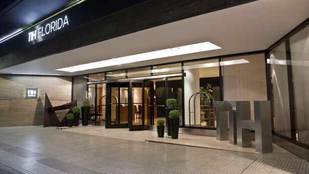 Entrada moderna de un hotel con fachada de vidrio, un cartel que dice "NH FLORIDA" y algunas plantas en macetas al frente.