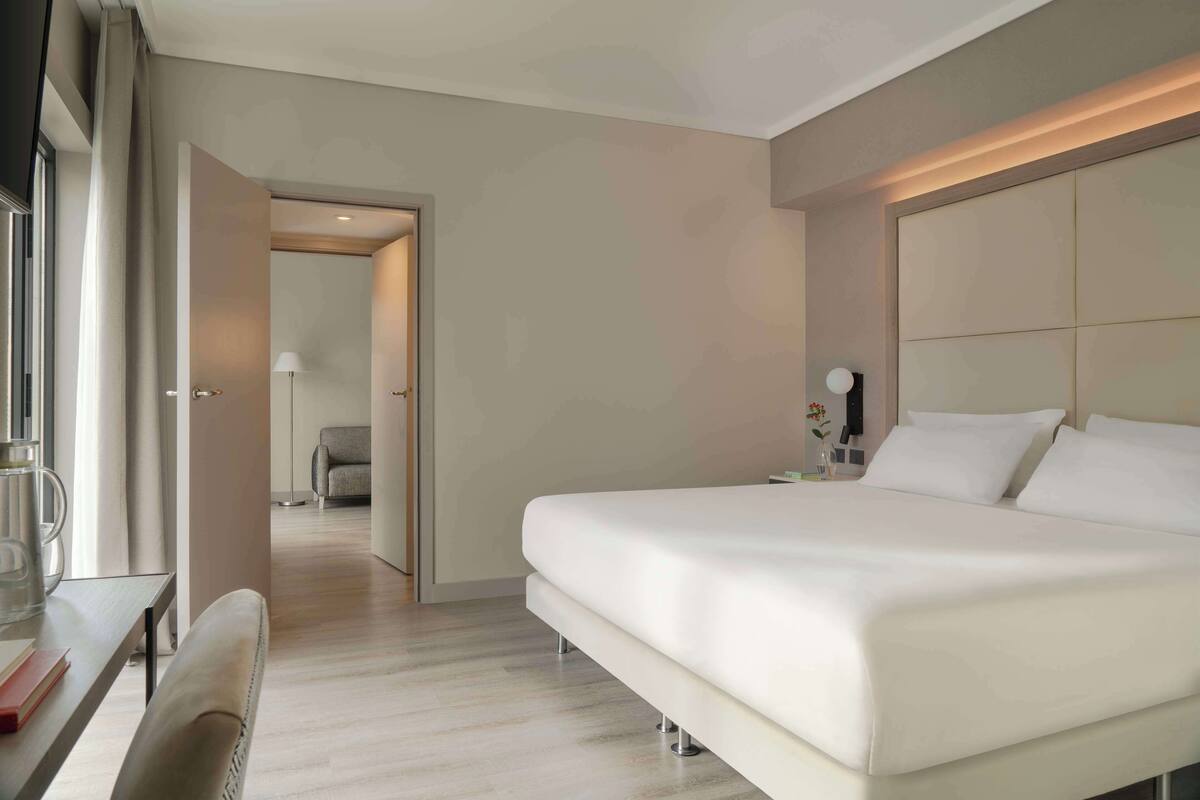 A modern hotel room with a large bed, a desk with a chair, and a view of the hallway.