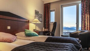 A hotel room with two beds, a desk, a chair, and a view of the mountains through the window. - Melbu Hotell  (Hadsel)