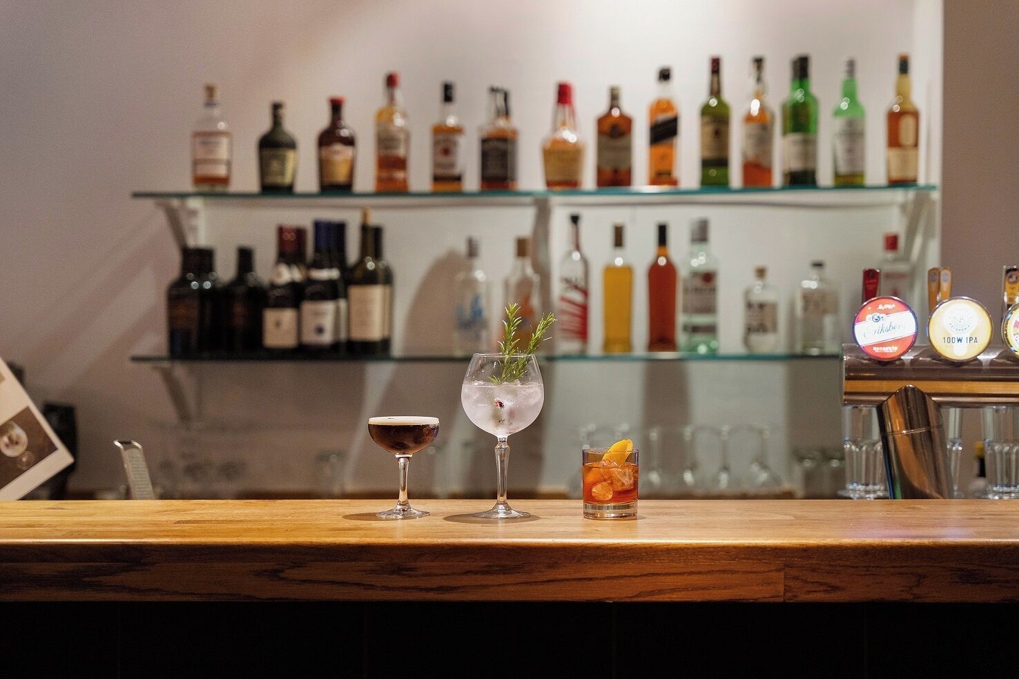 A bar area with a variety of liquor bottles on shelves and three glasses of different drinks on the counter.