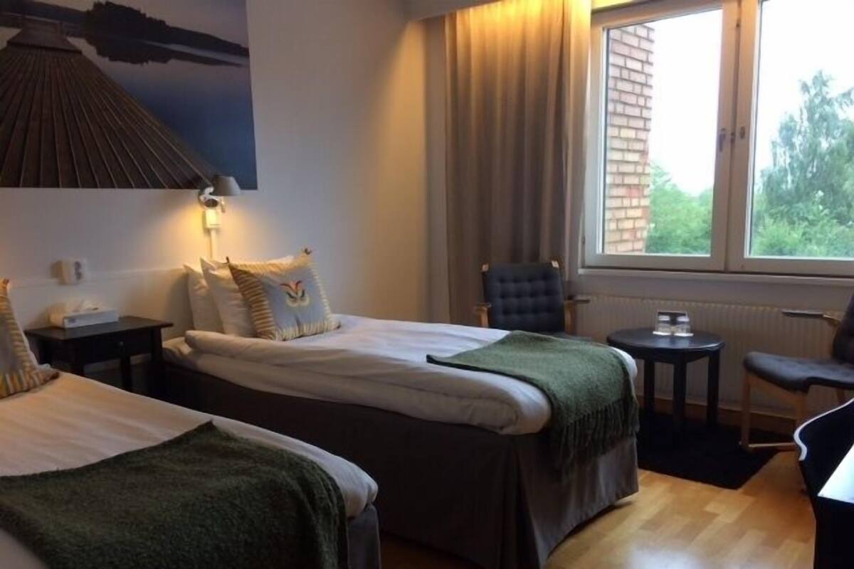 Habitación de hotel con dos camas, una silla, una mesita, una lámpara y una ventana con vistas a zonas verdes.