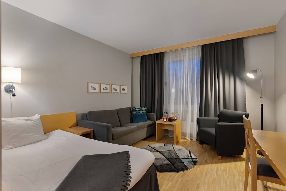 Habitación de hotel con cama, sofá, escritorio y silla. Hay una ventana con cortinas, una lámpara y cuadros en la pared.