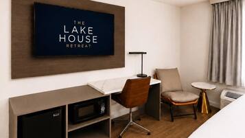 A hotel room with a flat-screen TV displaying 'The Lake House Retreat' on the wall, a desk with a chair, a small table with a lamp, a microwave, and a bed.
