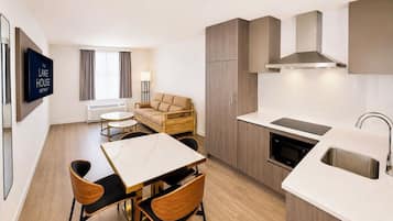 Une chambre d’hôtel moderne équipée d’une table à manger, de chaises, d’un canapé, d’une télévision, d’un coin cuisine et d’une fenêtre avec des rideaux.