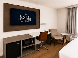 Habitación de hotel con televisor de pantalla plana, escritorio con silla, mesita de noche con lámpara, cama y ventana con cortinas.