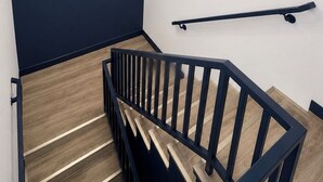 Una escalera con barandilla negra, peldaños de madera, una lámpara fijada a la pared y un pasamanos a la derecha.