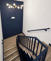 Una escalera con barandilla negra, peldaños de madera, una lámpara fijada a la pared y un pasamanos a la derecha.