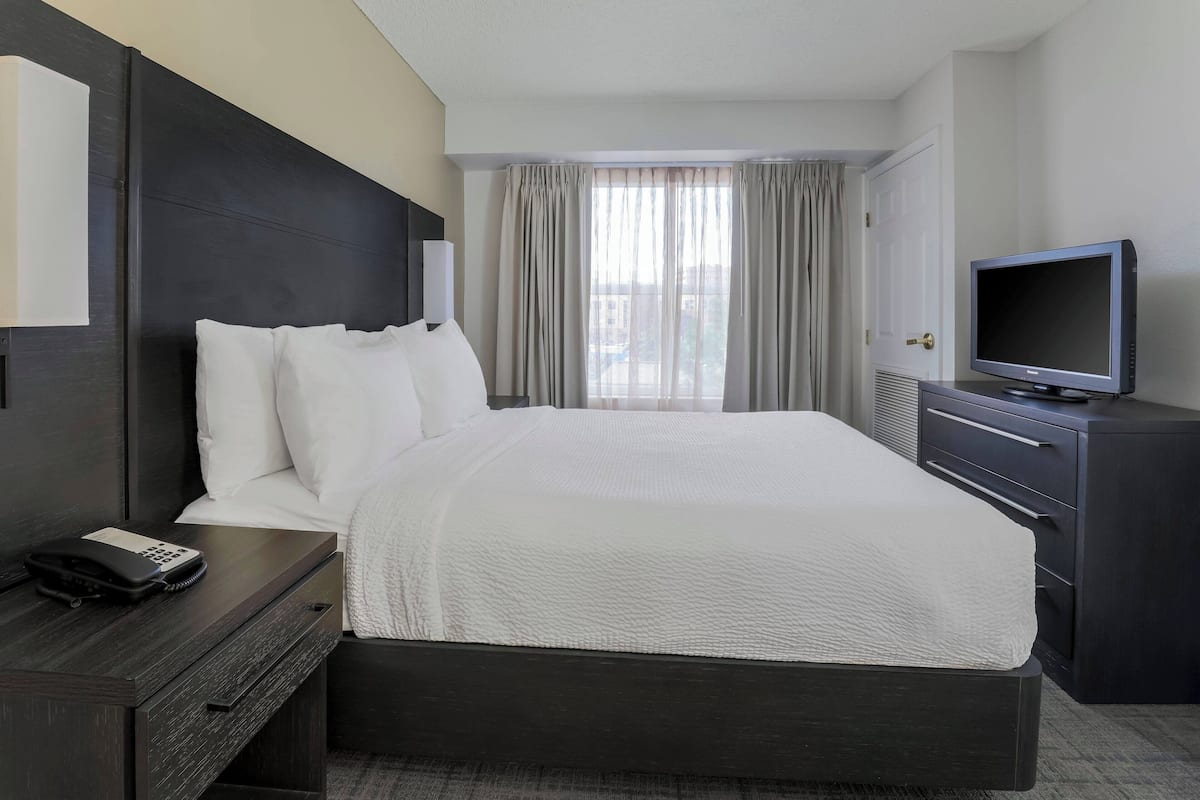 A hotel room with a large bed, a bedside table with a telephone, a television on a stand, and a window with curtains.