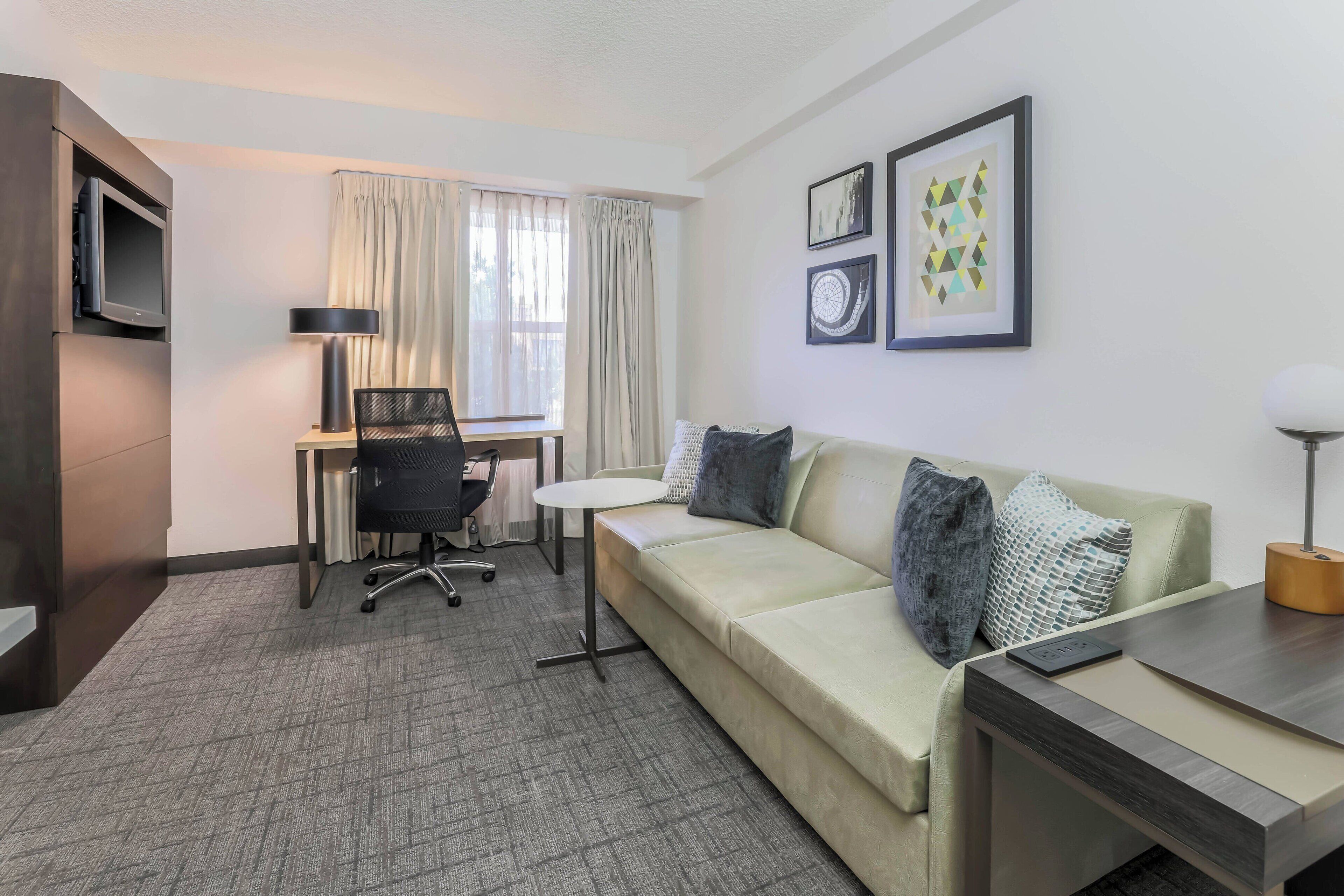 A hotel room with a sofa, a desk, a chair, a TV, and a lamp.