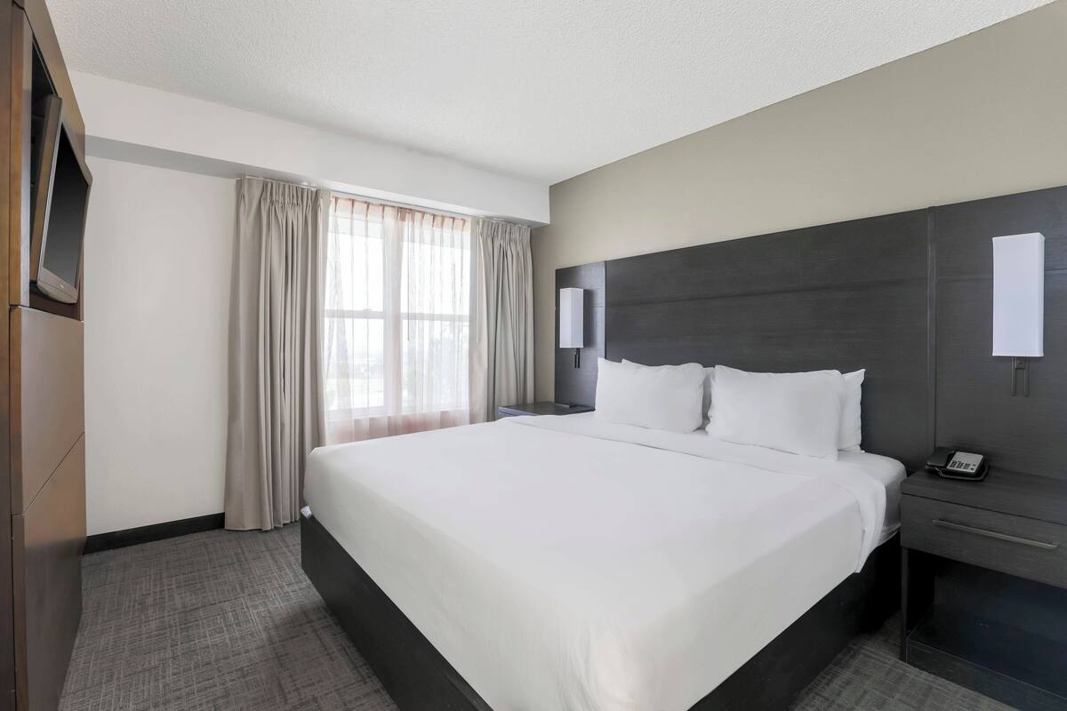 A hotel room with a large bed, a bedside table, a flat-screen TV, and a window with curtains.
