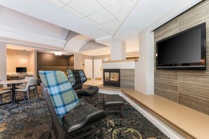 A hotel room with a flat-screen TV mounted on the wall, a fireplace, leather seating, a dining area with a table and chairs, and a patterned carpet.