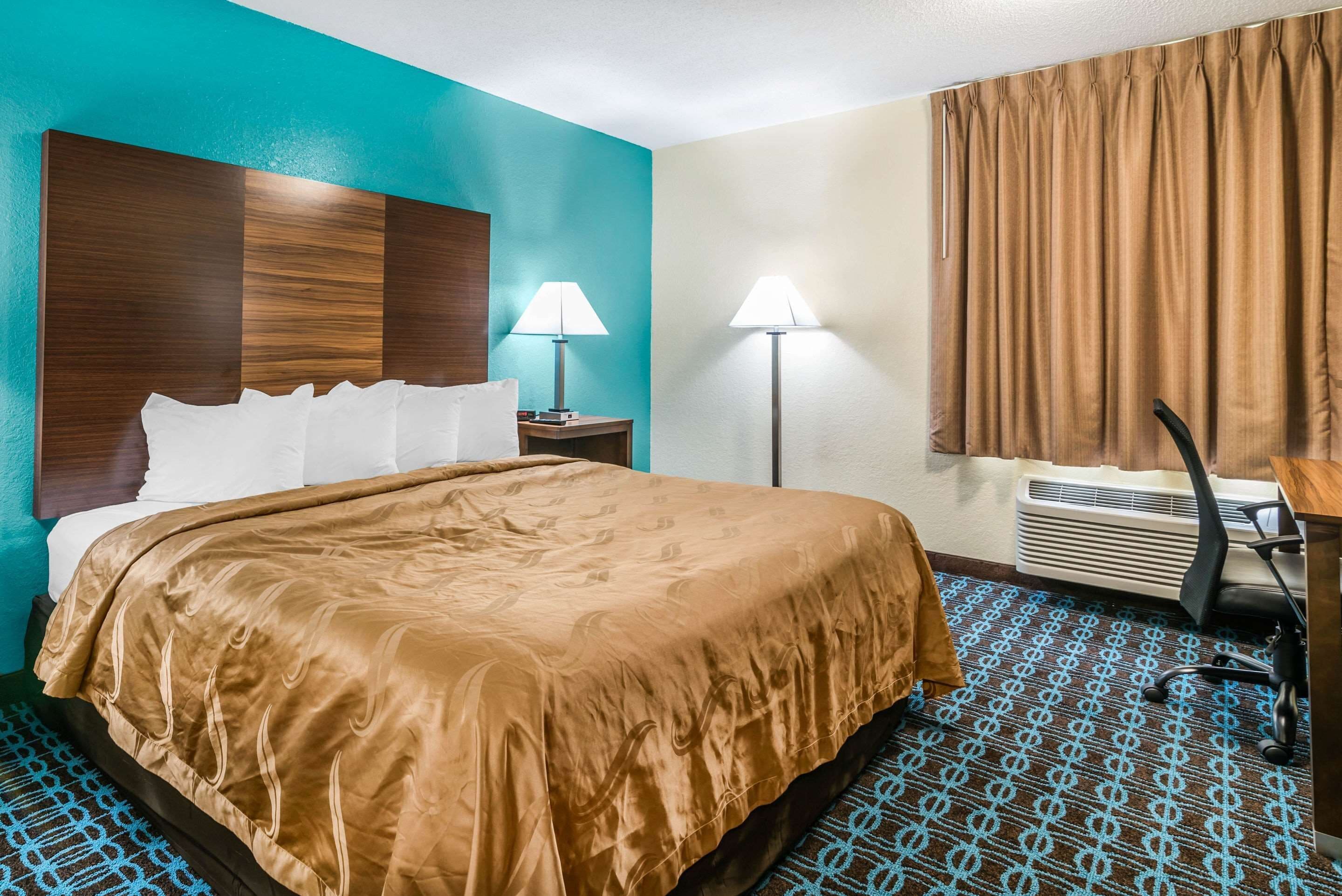 A hotel room with a bed, two bedside lamps, a desk with a chair, and a window with curtains.