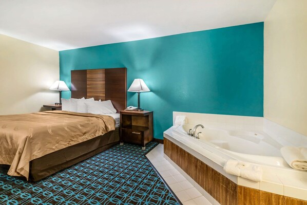 A hotel room with a bed, two bedside tables with lamps, a large bathtub, and a patterned rug.
