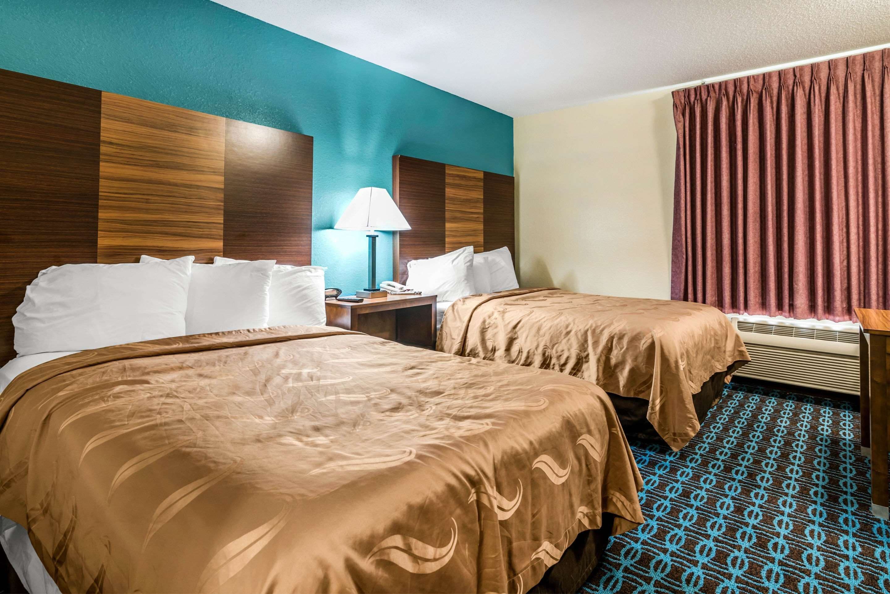 A hotel room with two beds, a nightstand, a lamp, and a window with curtains.