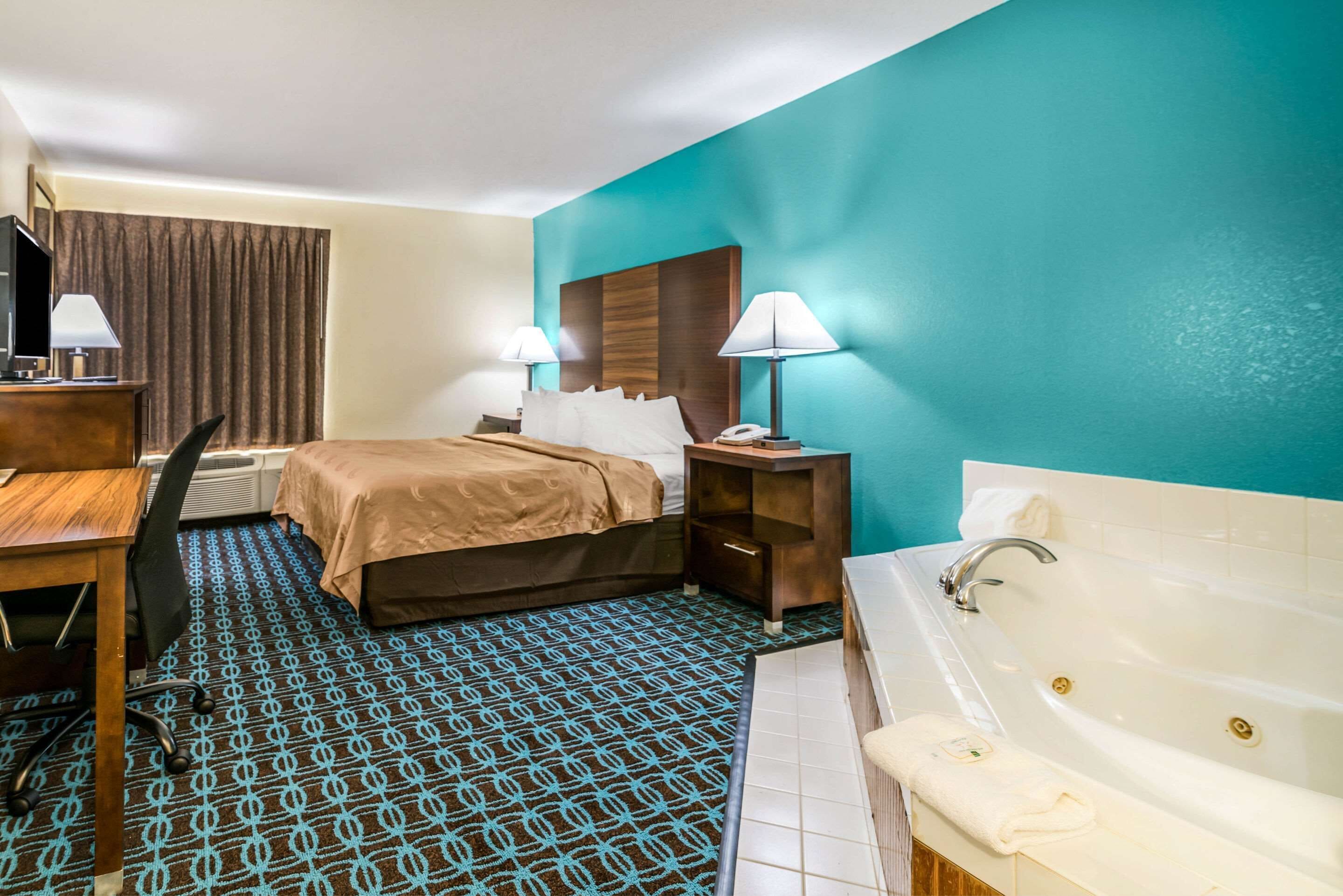 A hotel room with a bed, a desk with a chair, a desk lamp, a telephone, a nightstand with a lamp, and a bathtub.