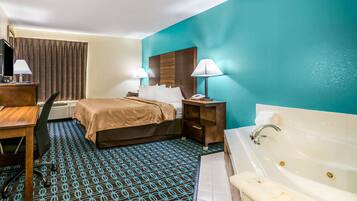A hotel room with a bed, a desk with a chair, a desk lamp, a telephone, a nightstand with a lamp, and a bathtub.