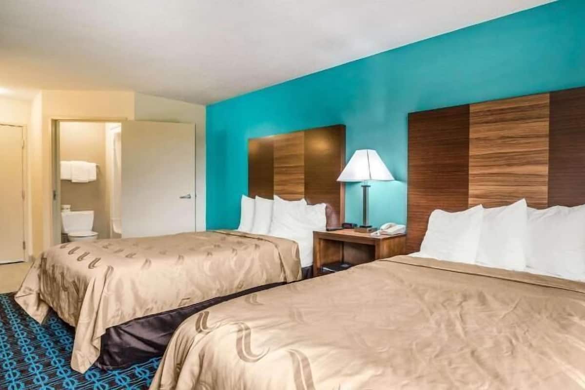 A hotel room with two beds, a nightstand with a lamp, and a bathroom visible in the background.