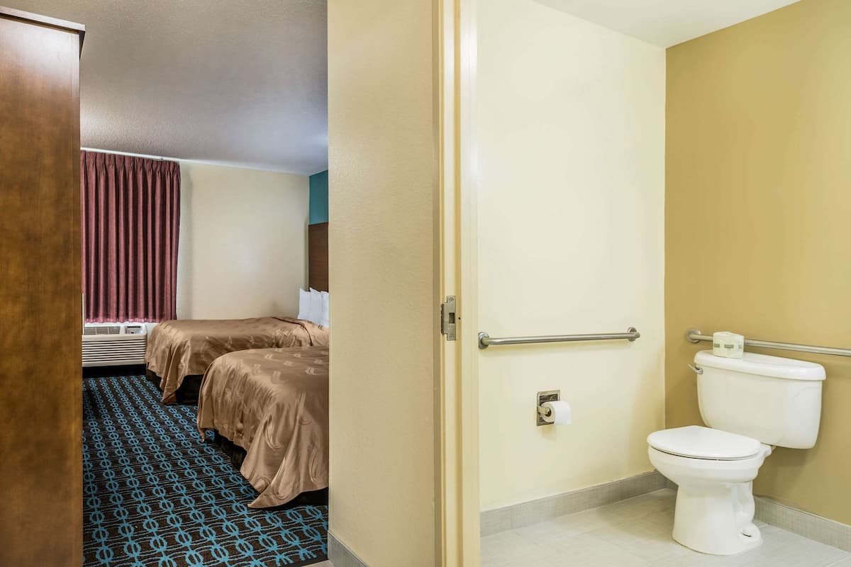 A hotel room with two beds, a bathroom with a toilet and handrail, and a patterned carpet.
