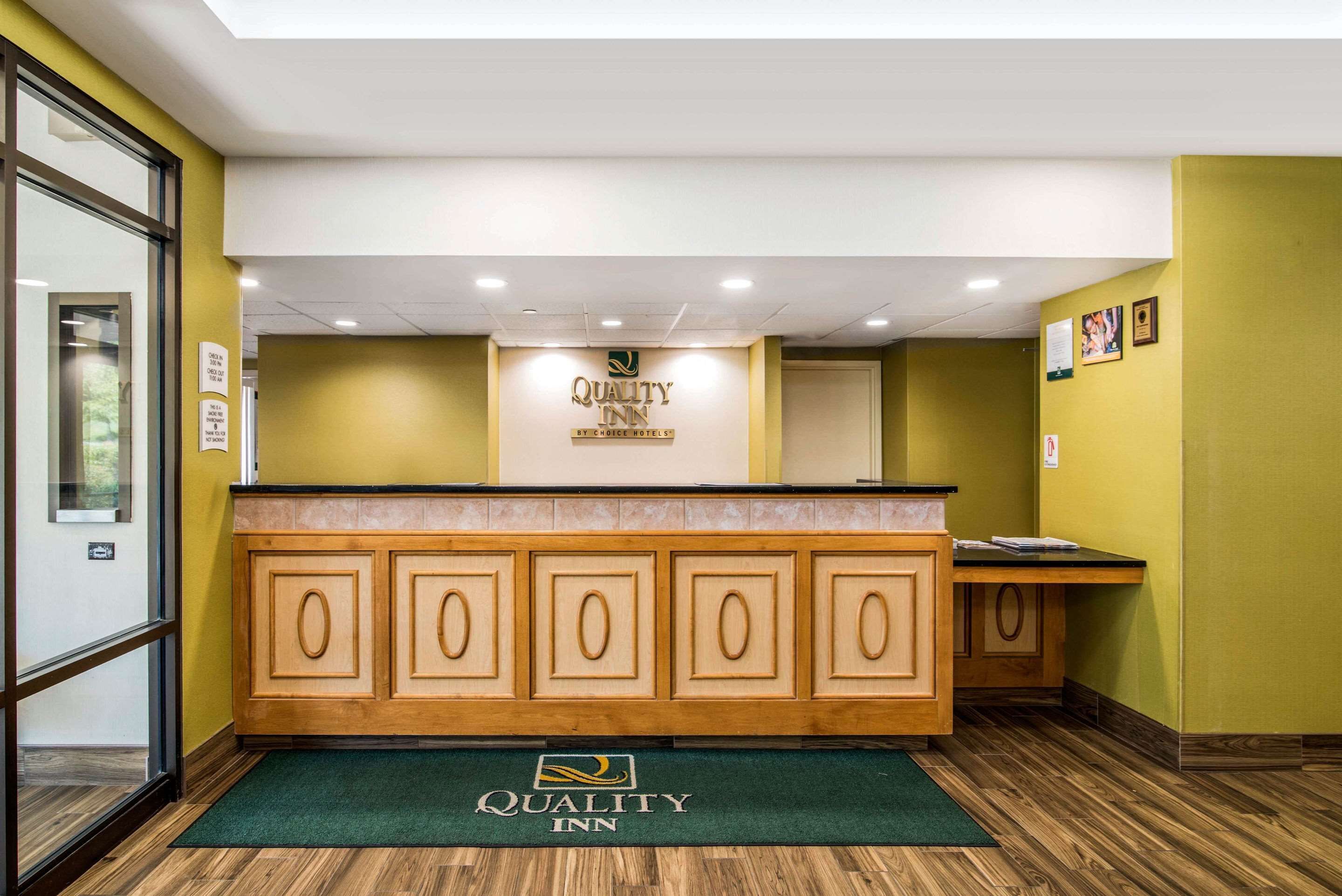 Una zona de recepció amb un taulell de fusta, un estora verd amb el logotip de l'hotel i un cartell sobre el taulell que diu "Quality Inn".