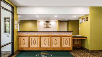 Un área de recepción con un mostrador de madera, un tapete verde con el logotipo del hotel y un cartel sobre el mostrador que dice "Quality Inn".