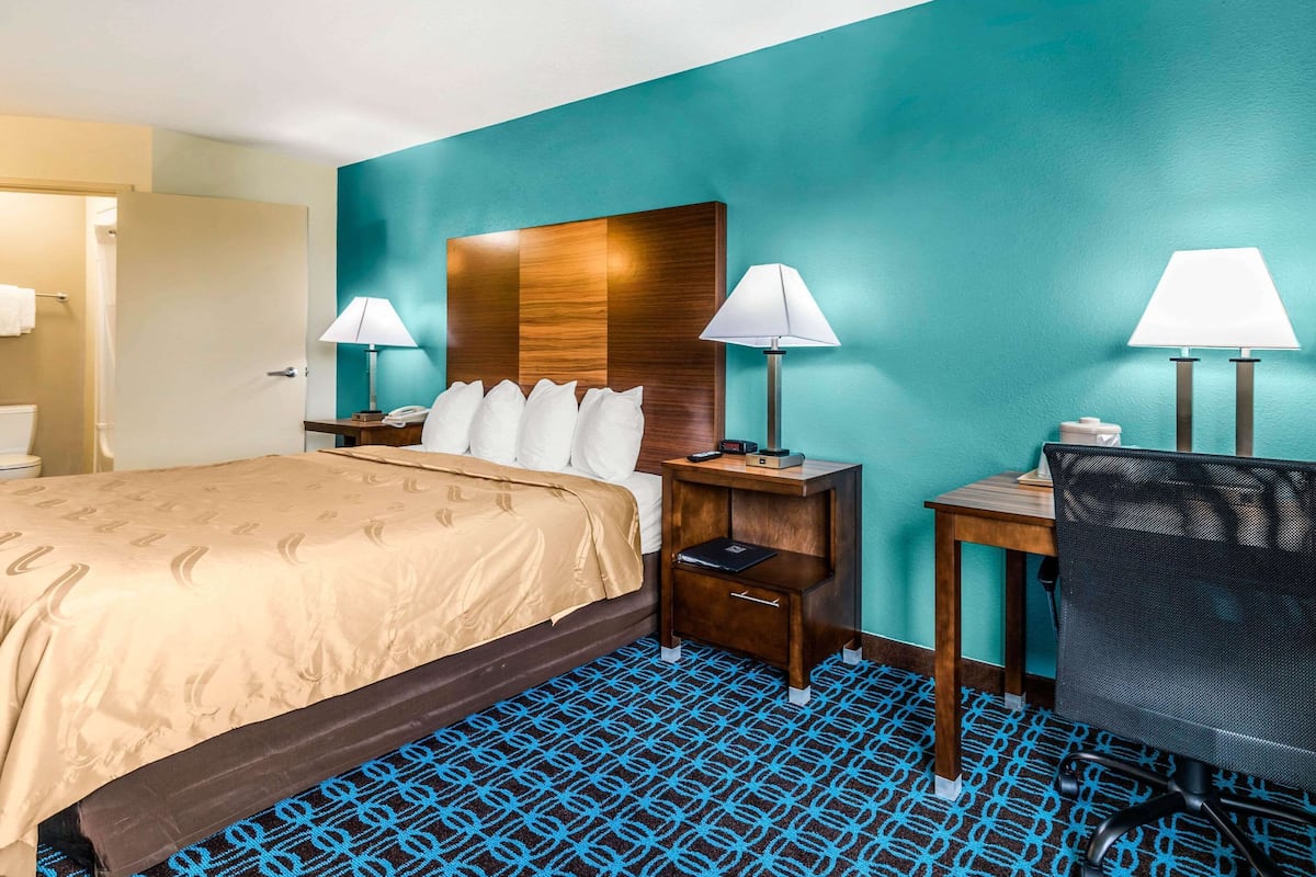 A hotel room with a bed, two bedside tables with lamps, a desk with a chair, and a patterned carpet.