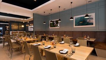 Ein modernes Restaurant mit langen Holztischen, beigen Stühlen und Pendelleuchten.