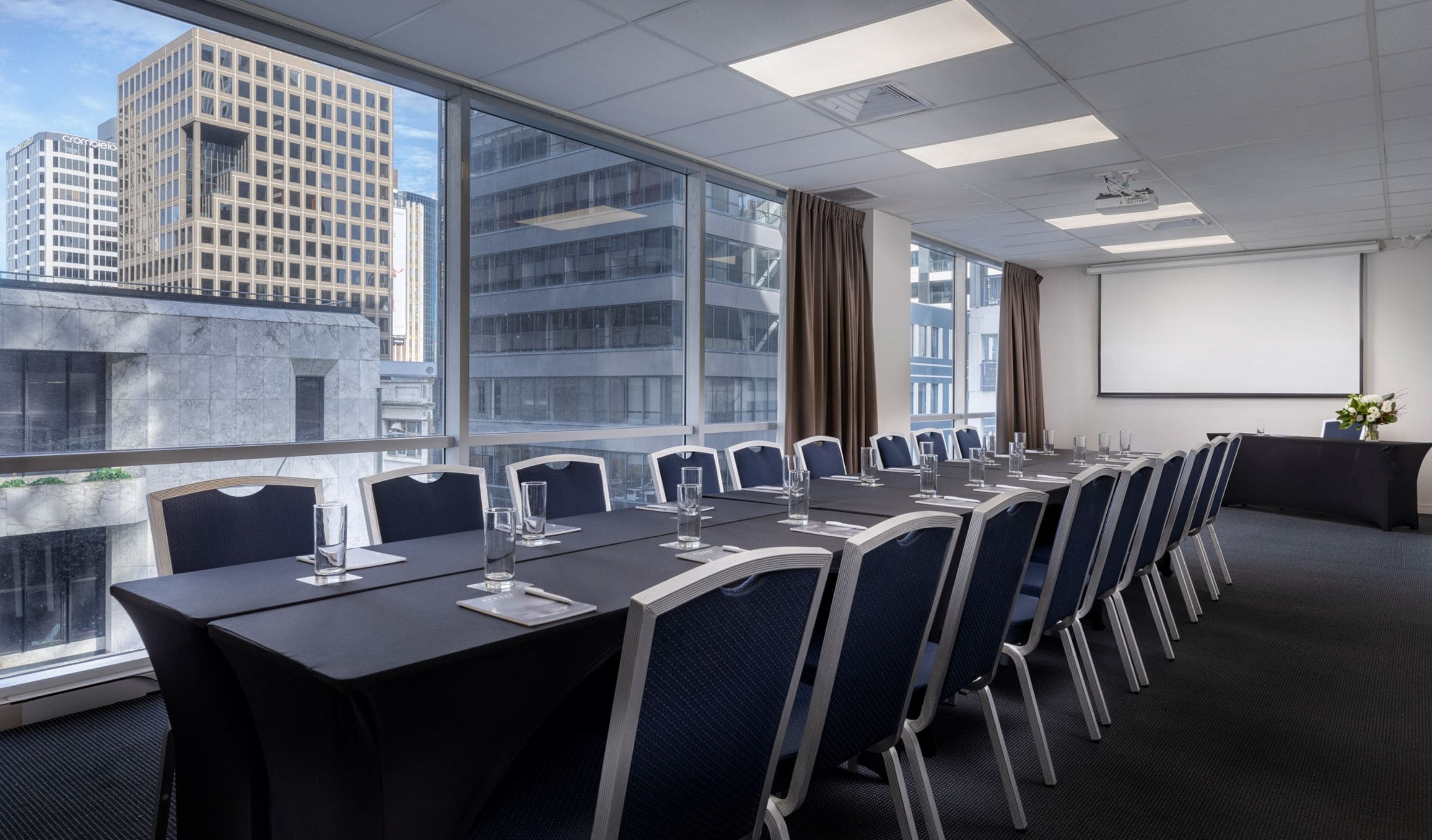 Uma sala de conferências com uma mesa comprida, cadeiras, um projetor e janelas amplas com vista para a cidade.