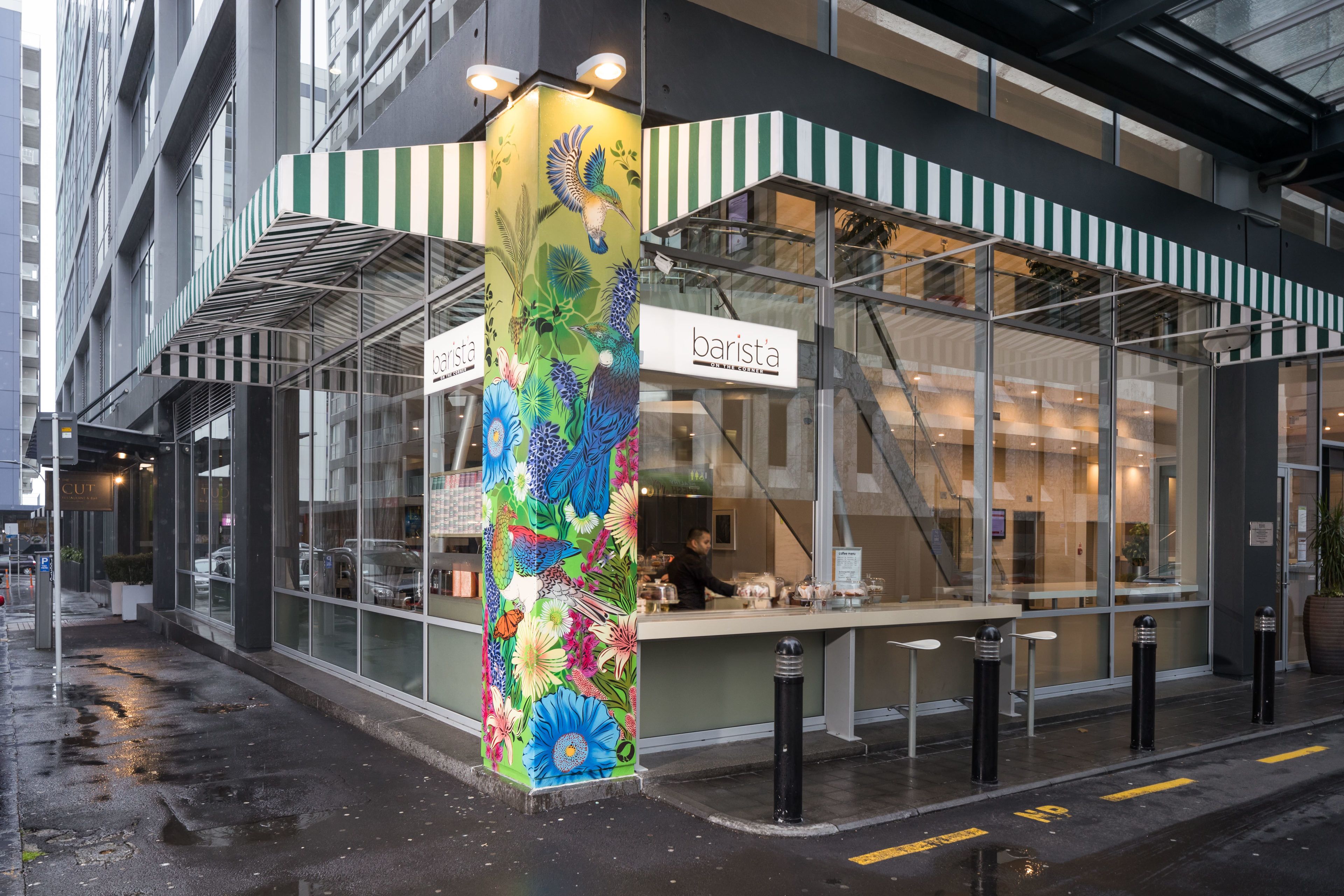 A modern barista shop with a colorful mural, glass storefront, and a staircase leading to an upper level.
