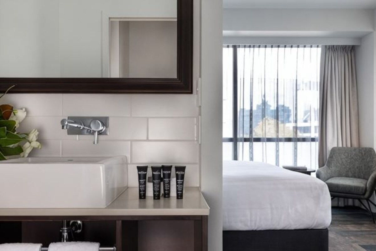 A hotel room with a white sink, mirror, toiletries, a bed, and a window with curtains.