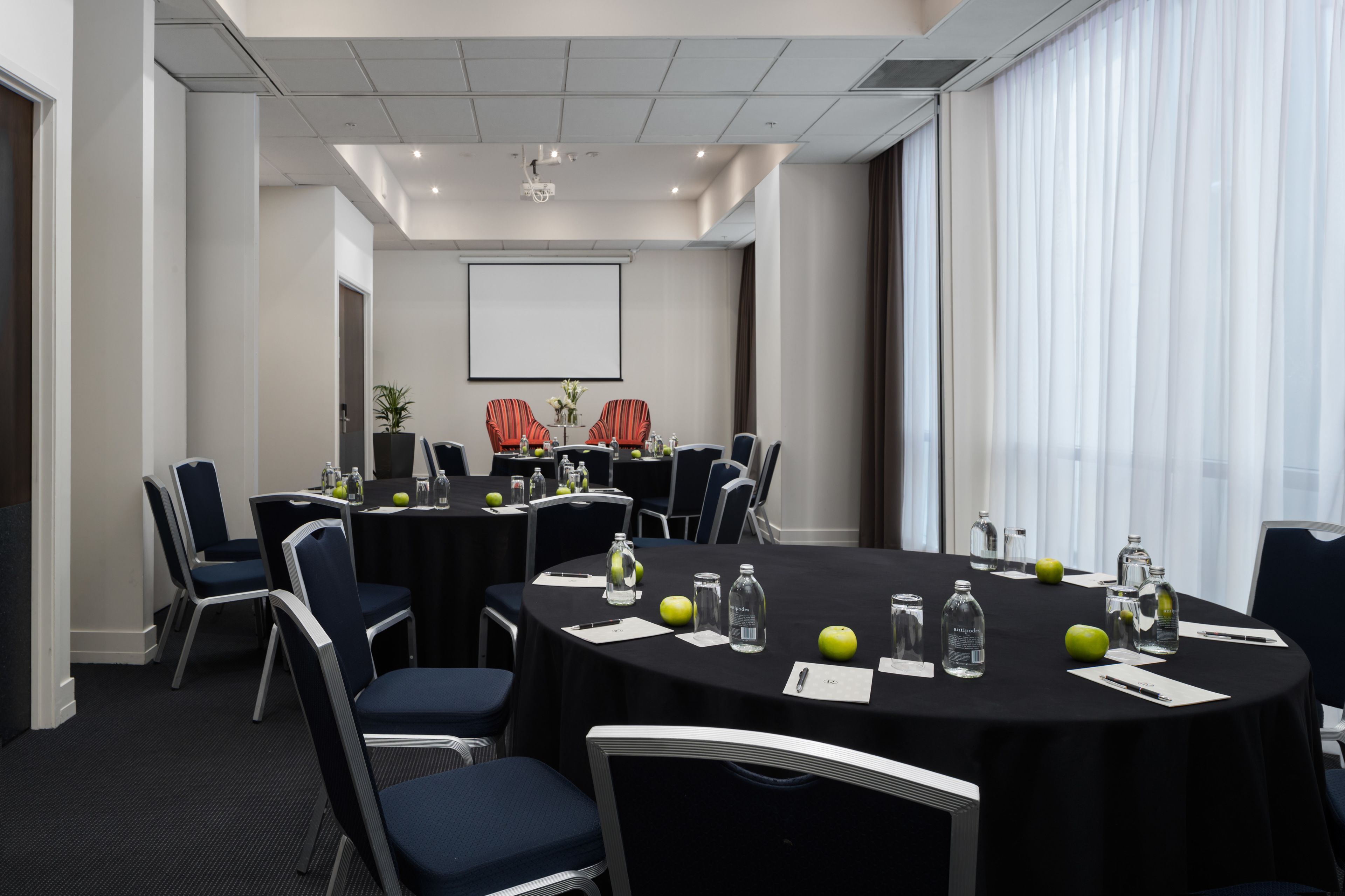 Uma sala de conferências com mesas redondas, cadeiras e uma tela de projetor.