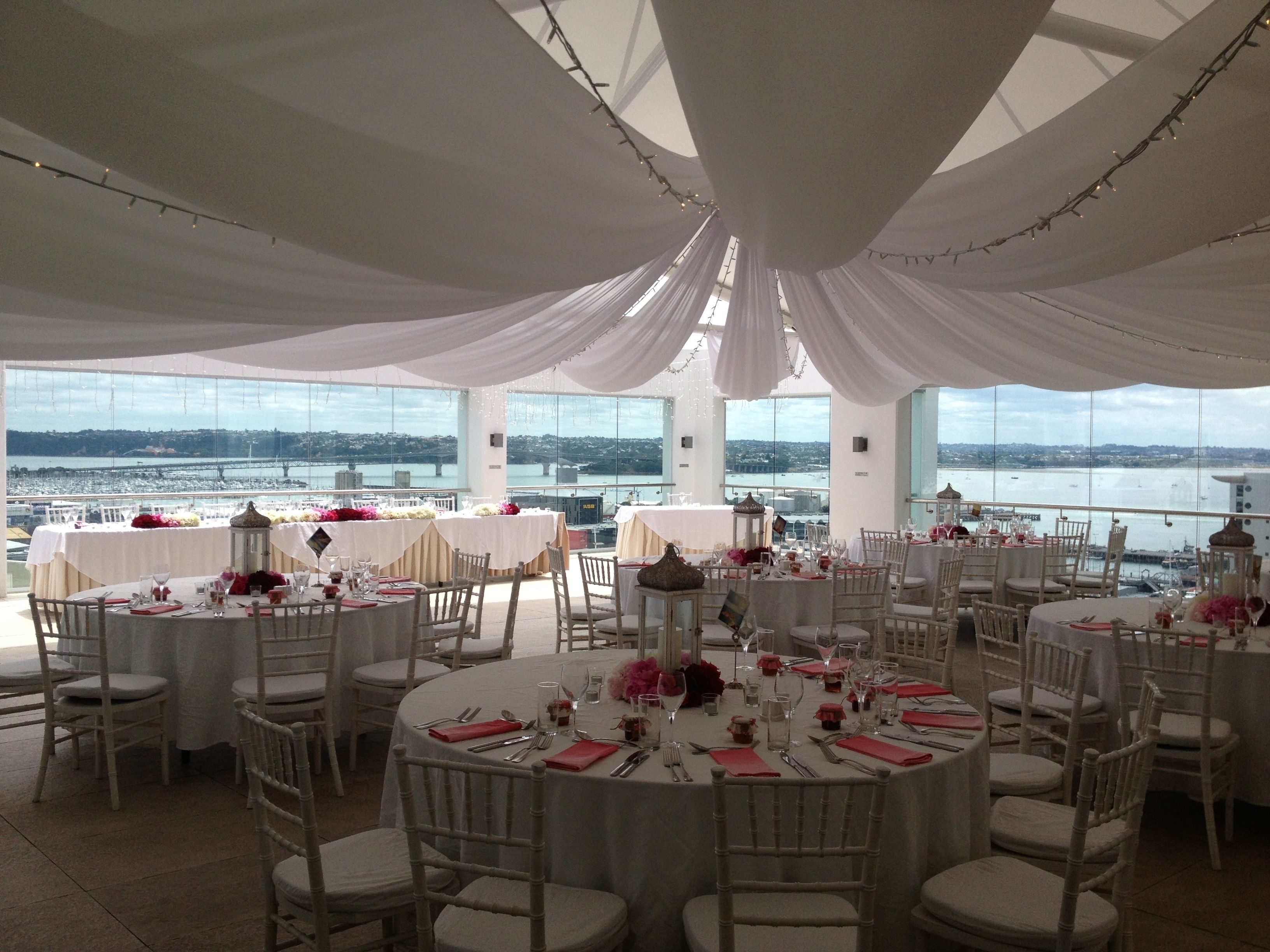 A banquet hall with round tables set for dining, large windows offering a view of the water and cityscape, and draped white fabric on the ceiling.