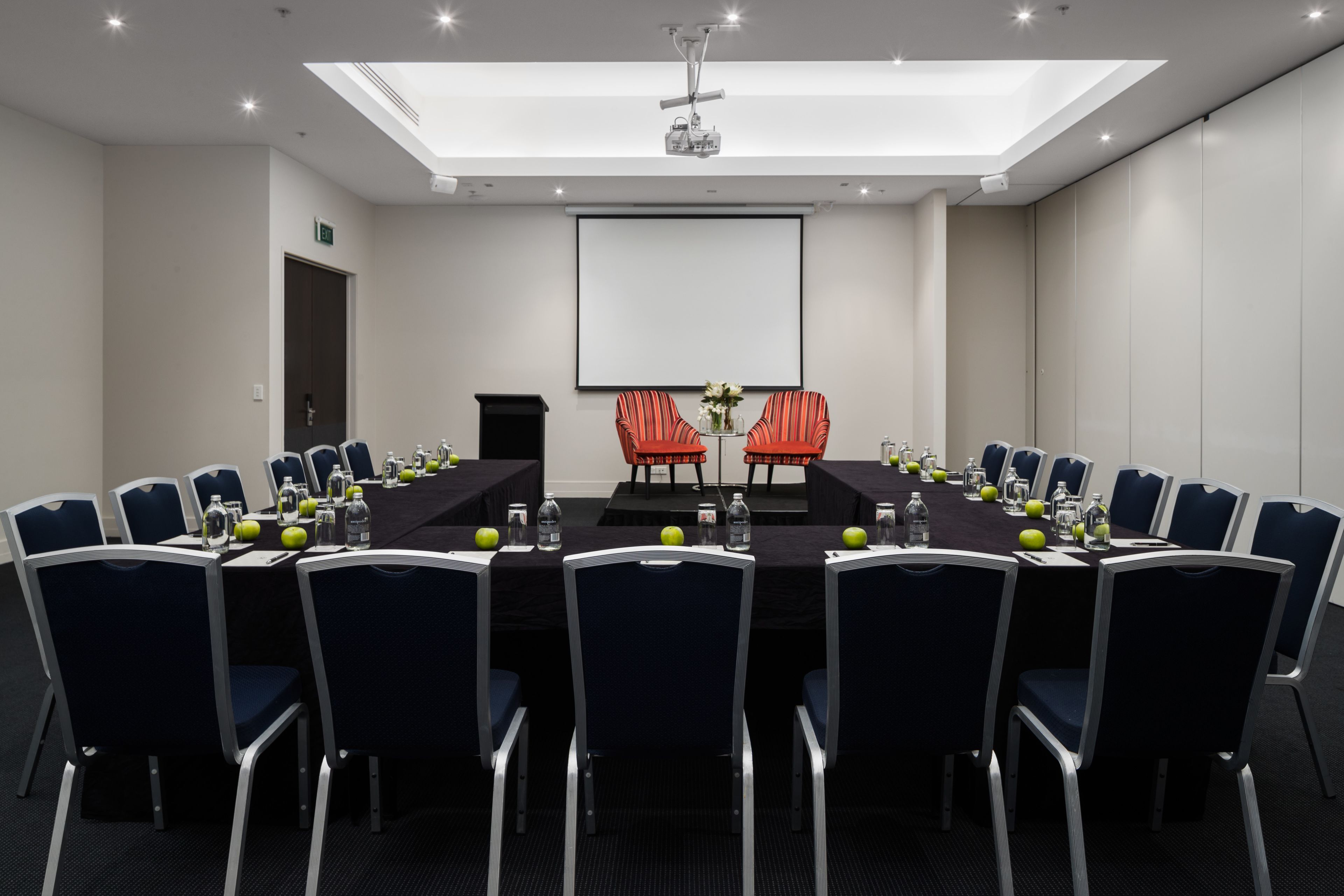 A conference room with a long table, chairs, a projector screen, and a speaker.