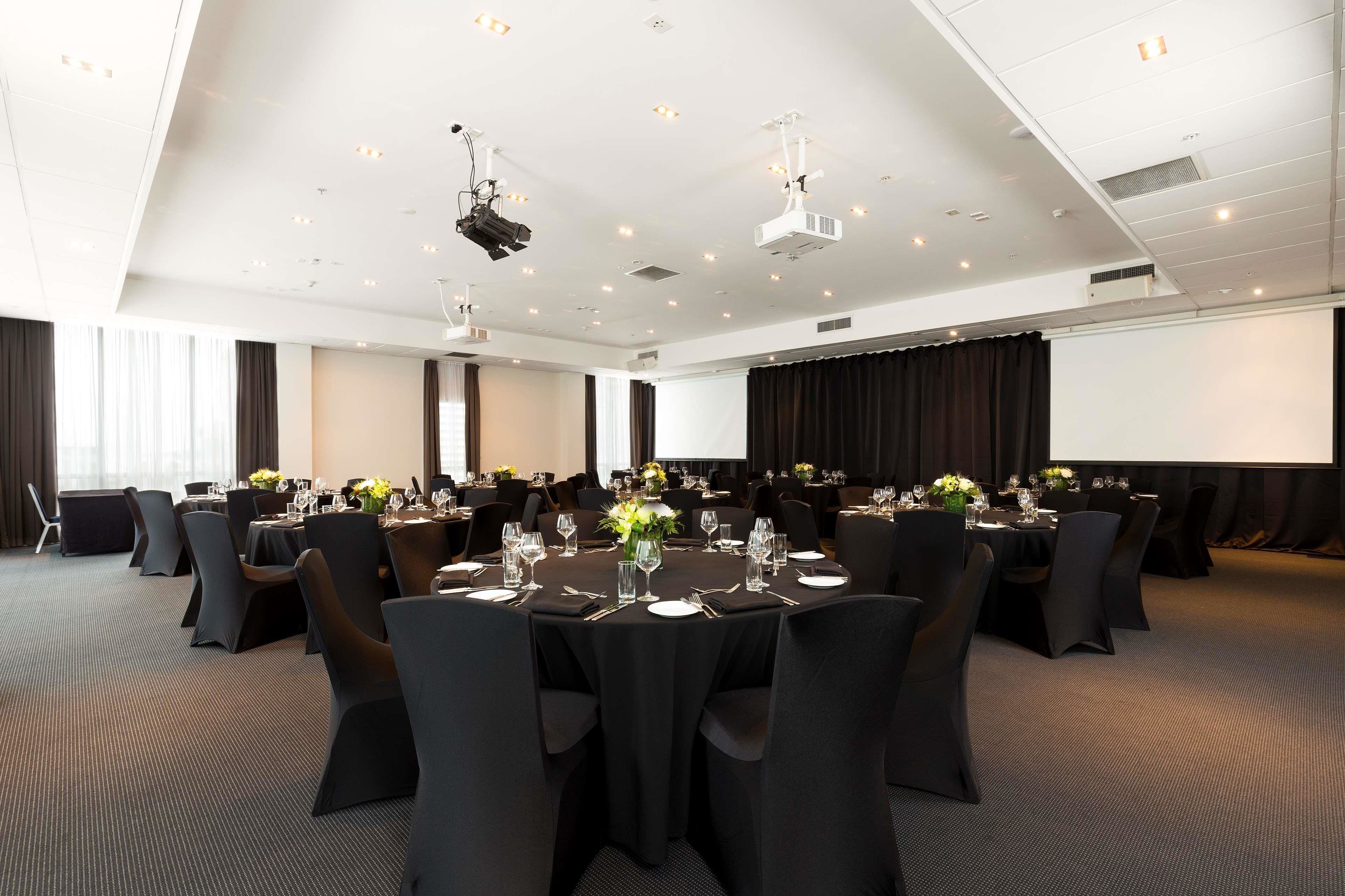 Sala de conferências com várias mesas preparadas para um evento formal, equipada com projetores e uma tela grande.