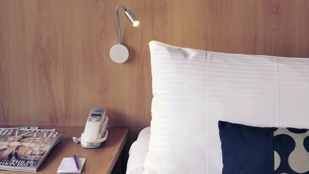 A neatly made bed with white and navy blue pillows, a wooden nightstand with a magazine, a telephone, and a wall-mounted lamp. - Mercure North Melbourne (North Melbourne)