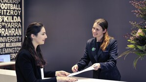 Deux personnes se tiennent derrière un comptoir d’accueil, et on peut voir, en arrière-plan, un panneau indiquant une liste de noms.