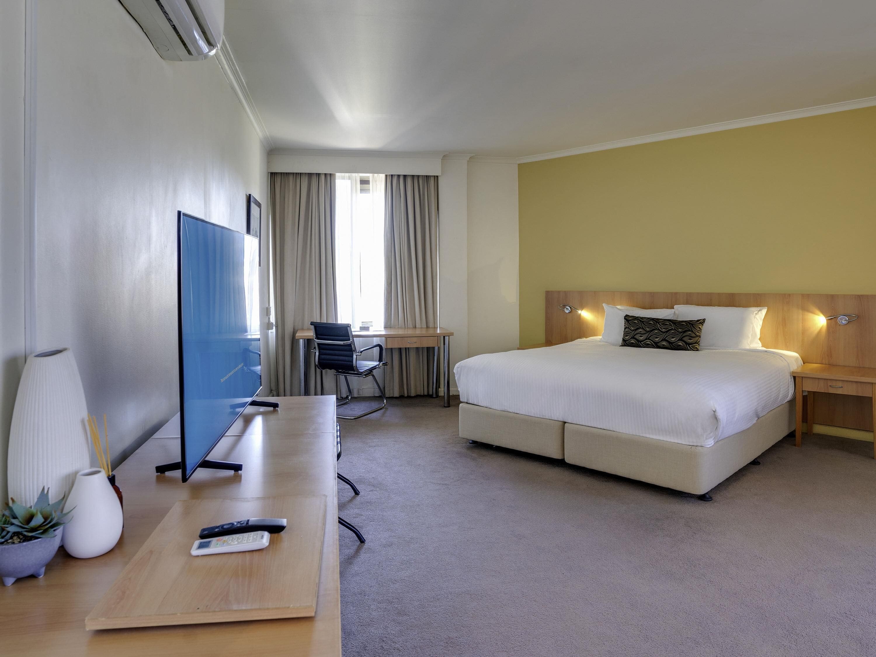 A hotel room with a bed, desk, chair, TV, and a small table with a remote control and phone.