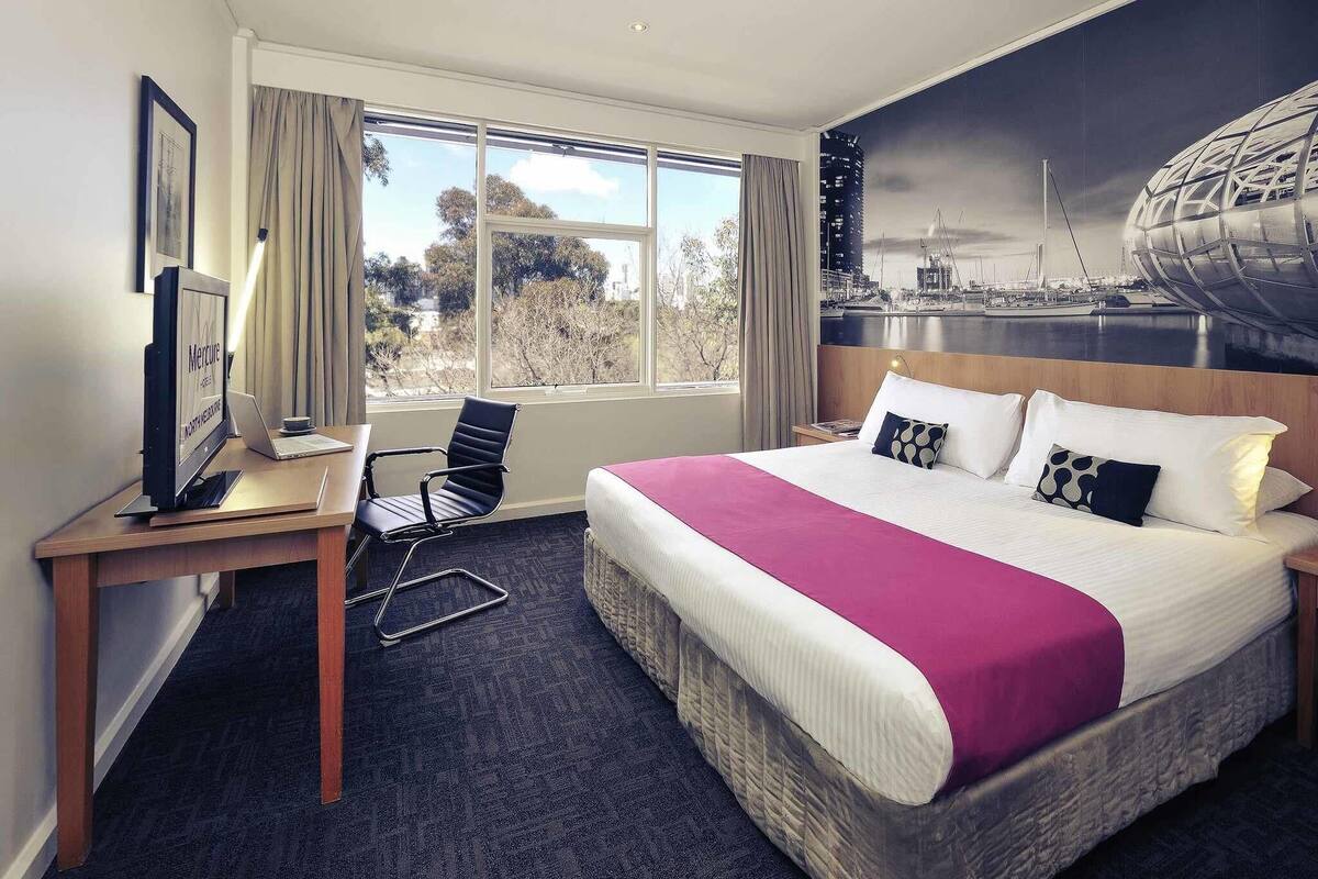 Habitación de hotel con cama, escritorio con silla, televisor y un amplio ventanal con vista a la ciudad.