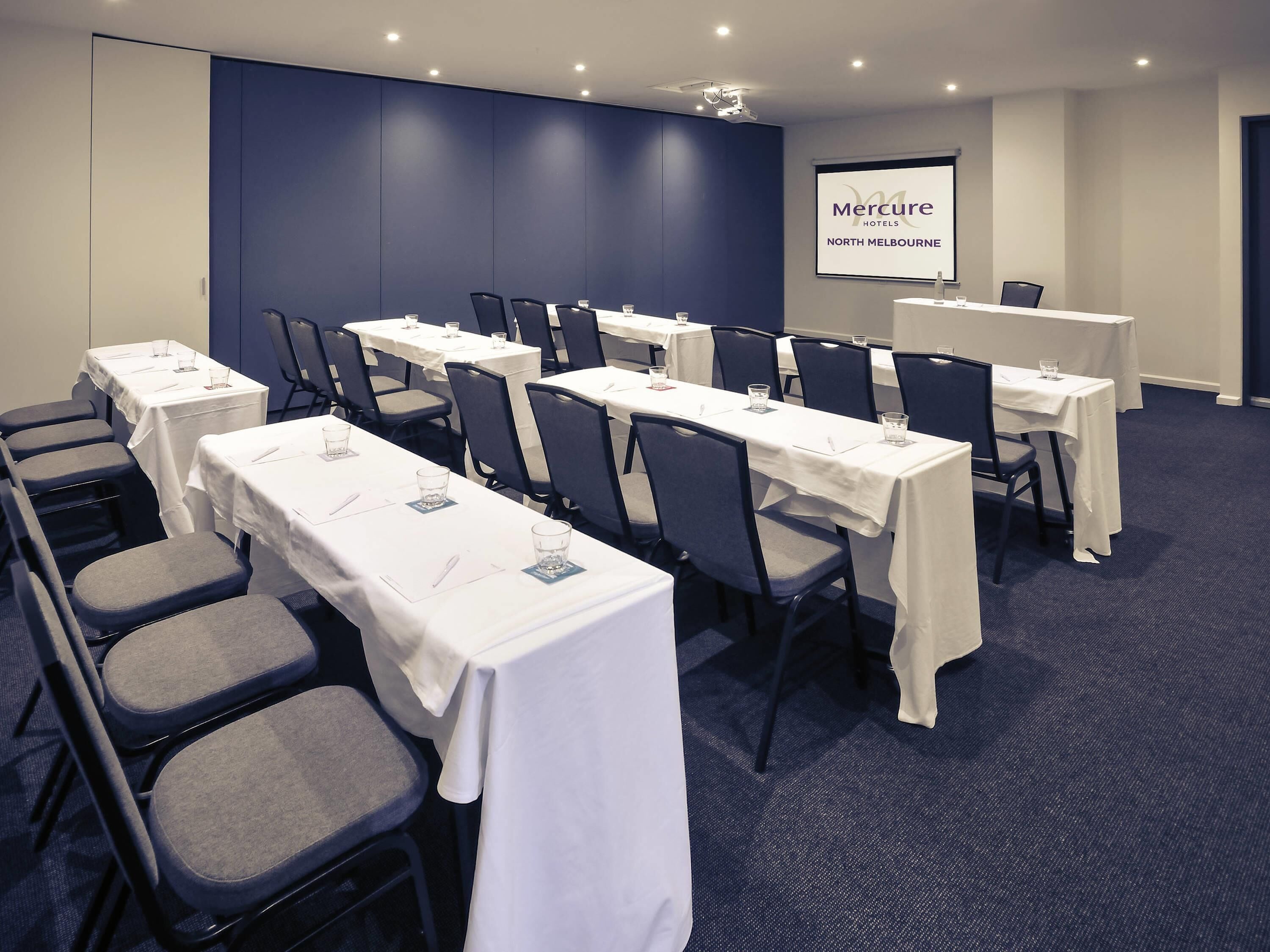 Ein Konferenzraum mit Tischen, Stühlen, einer Projektionsleinwand und einem Schild für Mercure Hotels North Melbourne.